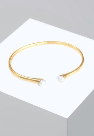 Guldmetallarmband i cuff-stil med en slät, rundad design som har två vita pärlprydnader i varje ände. Minimalistiskt och elegant utseende.
