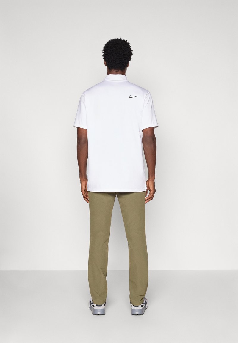 Witte polo shirt met een zwart Nike-logo op de achterkant, gecombineerd met olijfgroene broek en grijze sneakers, gezien vanuit de achterkant.