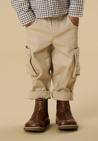 Beige corduroy pants with rolled cuffs and side pockets, paired with brown leather ankle boots. The shirt features a brown and white checkered pattern.