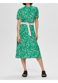 Vestito floreale verde con maniche corte, colletto a bottoni e una cintura bianca; lunghezza al ginocchio con orlo fluente. Indossato con sandali neri.