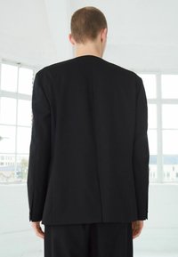 Sacou negru, croi elegant, cu o textură fină, fanteză la spate și mâneci de lungime medie, expus pe un fundal luminos și aerisit.