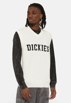 Chaleco de punto blanco con escote en forma de V, que presenta la palabra "DICKIES" en letras negras en negrita. Lleva una camiseta de manga larga negra debajo.