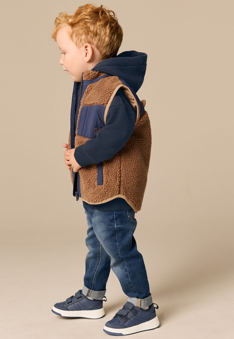 Jeune enfant aux cheveux roux bouclés portant un sweat à capuche bleu, un gilet en polaire marron, un jean retroussé et des baskets bleu marine, debout de profil sur un fond beige.
