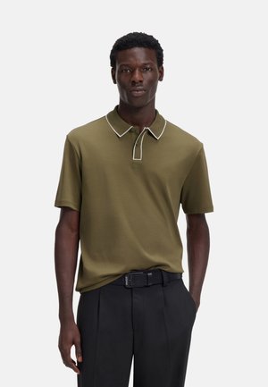 Hombre con camisa polo verde oliva con ribete blanco y pantalones negros, de pie con una mano en el bolsillo frente a un fondo liso.