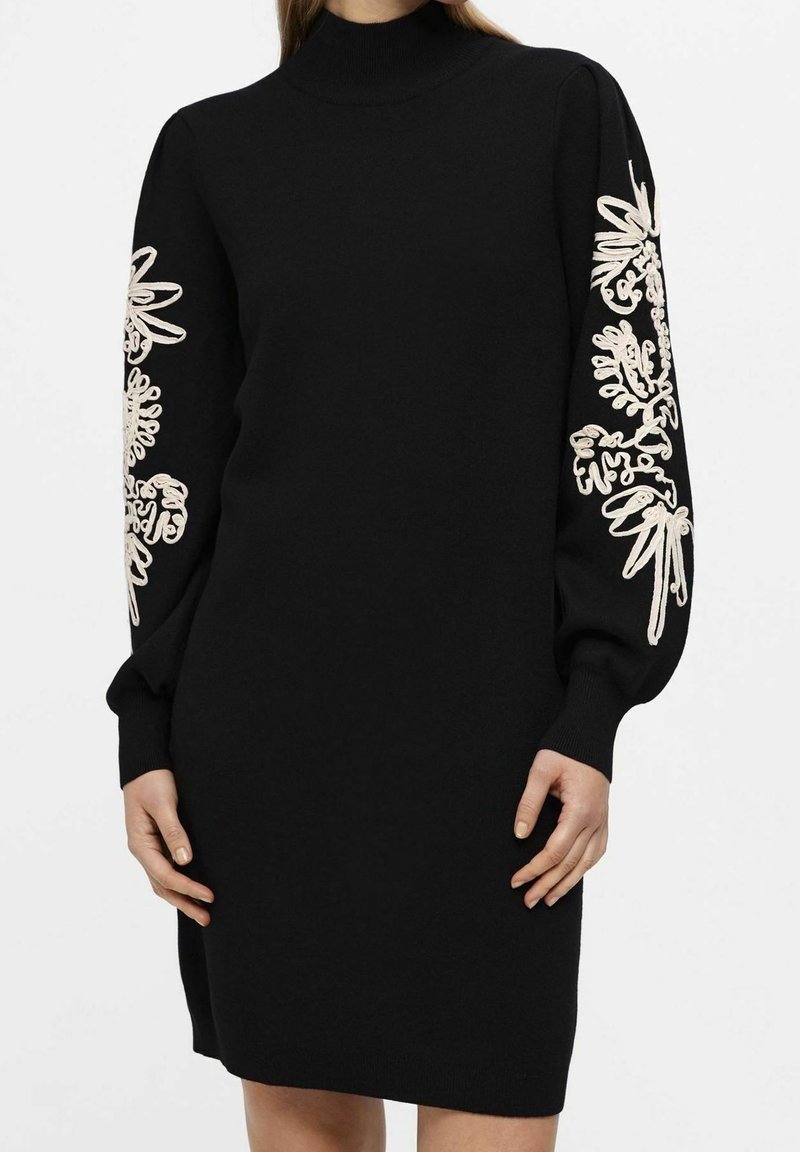 Robe noire à manches longues avec col montant et broderie florale blanche le long des manches, portée par une femme debout, les mains relâchées le long du corps.