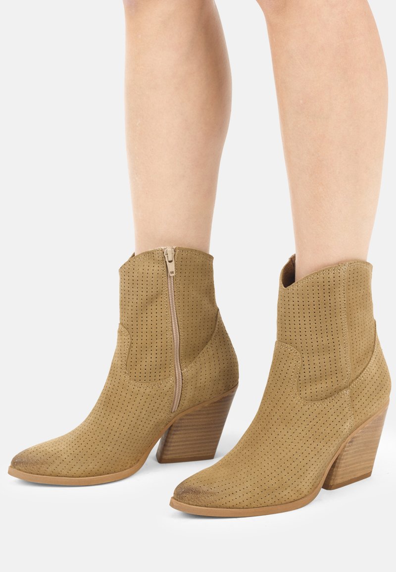 Bottines en daim perforé couleur beige avec fermeture éclair latérale et talons blocs en bois superposés, portées sur jambes nues.