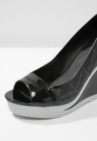 Svart lack wedge sandal med öppen tå, med Calvin Klein-logotypen på sidan och en vit gummisula. Enkelt och stilrent design.