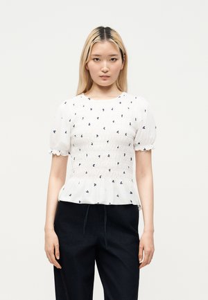 Jeune femme aux cheveux blonds raides portant un chemisier blanc à manches courtes avec des motifs floraux noirs et un pantalon noir sur un fond uni.