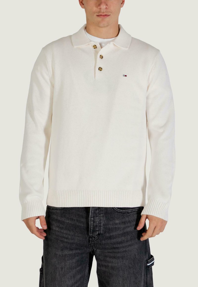 Biały sweter polo z dzianiny z prążkowanymi mankietami i krawędzią, z trzema guzikami i małym logo na piersi. Noszony z ciemnymi dżinsami.
