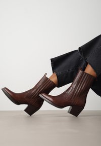 Botas de cuero marrón hasta el tobillo con un diseño texturizado y acanalado, tacones cuadrados y costuras de acento, combinadas con pantalones de mezclilla negros deshilachados.