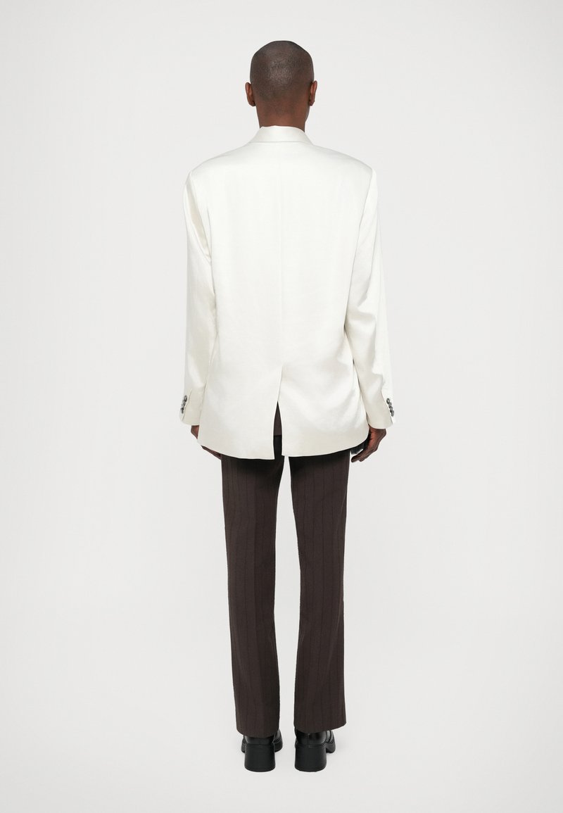 Vit skräddarsydd kavaj med en vent bak, parat med mörkbruna randiga byxor och svarta skor, sedd bakifrån.