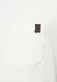 Λευκό βαμβακερό T-shirt με τσέπη που φέρει μαύρη ετικέτα Jack & Jones. Μαλακή υφή με κανονική εφαρμογή και καθαρό, μινιμαλιστικό σχέδιο.