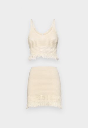 Strikket to-delt sett som består av en kort topp med en scalloped frynsehem og et matchende skjørt med lignende tekstur og frynse detaljer, begge i kremfarge.