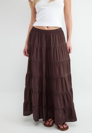 Kobieta ubrana w biały top bez rękawów i długą, warstwową ciemnobrązową spódnicę z sandałami na jednolitym tle.