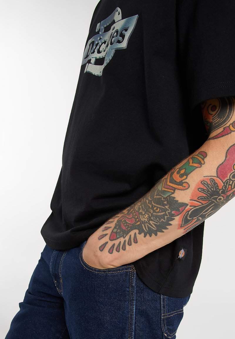 Black cotton t-shirt featuring a silver graphic logo on the chest, paired with dark blue denim jeans with visible stitching and a pocket.