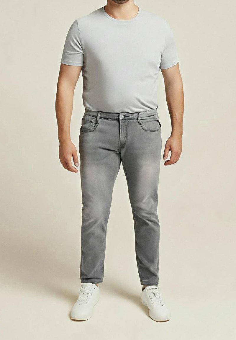 Homme portant un t-shirt ajusté gris clair, un jean gris et des baskets blanches, debout devant un fond clair uni.