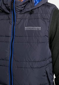 Marineblauer Steppweste mit blauen Akzenten, verfügt über eine Kapuze, einen Reißverschluss vorne und Logodetails. Hergestellt aus glattem, leichtem Material.