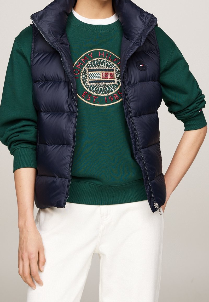 Marineblaue Steppweste mit hohem Kragen über grünem Sweatshirt mit gesticktem Logo und gerippten Bündchen, kombiniert mit weißen Hosen.