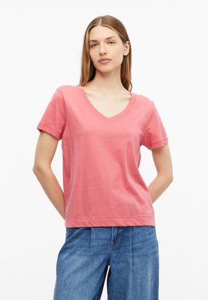 Młoda kobieta z prostymi włosami, ubrana w różowy T-shirt z dekoltem w serek i krótkim rękawem oraz niebieskie jeansy, stoi ze złożonymi rękami za plecami.