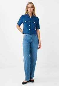 Junge Frau in einem kurzärmeligen blauen Knopf-Cardigan, hoch taillierten blauen Jeans und schwarzen flachen Schuhen, die vor einem weißen Hintergrund steht.
