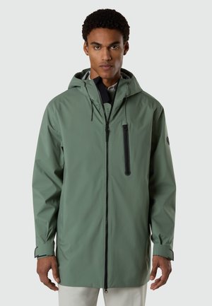 Joven con chaqueta impermeable verde con capucha, cremallera frontal y bolsillo con cremallera en el pecho, posando sobre un fondo liso.