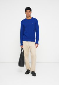 Kék kötött pulóver bordás mandzsettákkal és aljjal, bézs nadrággal párosítva. A modell egy fekete táskát tart, háttérben egy sima fehér felülettel.