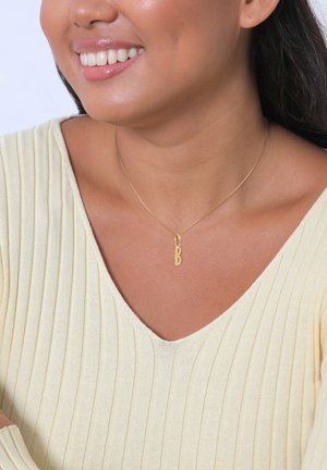 Goldkette mit einem strukturierten "B"-Anhänger, getragen zu einem gerippten, hellgelben Oberteil. Glatte Hautfarbe ist unter dem Ausschnitt sichtbar.