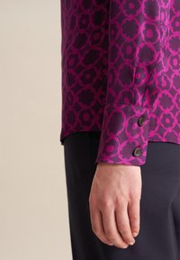 Camicia viola con motivo floreale, caratterizzata da un polsino bottonato e una texture liscia. Indossata con pantaloni scuri.