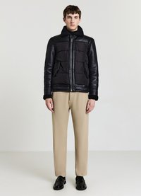 Veste noire matelassée avec manches en cuir et col en peau de mouton, associée à un pantalon beige et des chaussures noires. Texture lisse sur l'ensemble.