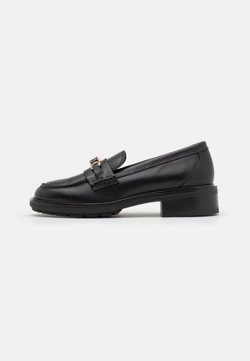 Mocassin noir en cuir avec petit talon, bout arrondi et détail boucle dorée à l'avant, présenté de profil sur un fond blanc.