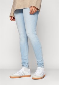 Vaqueros skinny azul claro con una rodilla desgastada, combinados con zapatillas blancas que tienen tres rayas grises y una suela de goma marrón.