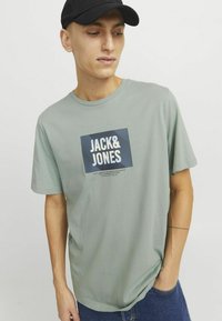 Svijetlozeleni pamučni majica s kratkim rukavima koja sadrži pravokutni grafički prikaz s natpisom "JACK&JONES" u debelom bijelom fontu, i opuštenim krojem.