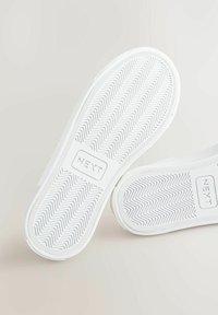 Tălpi albe de pantofi cu un model de zgârietură în zigzag, fiecare talpă având cuvântul "NEXT" într-o inserție rectangulară la mijloc.