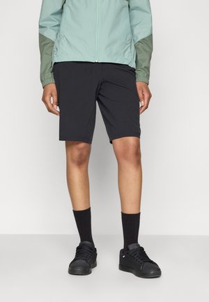 Personne portant un short noir mi-cuisses, des chaussettes noires, des baskets noires et une veste vert clair avec des accents de manches vert foncé.