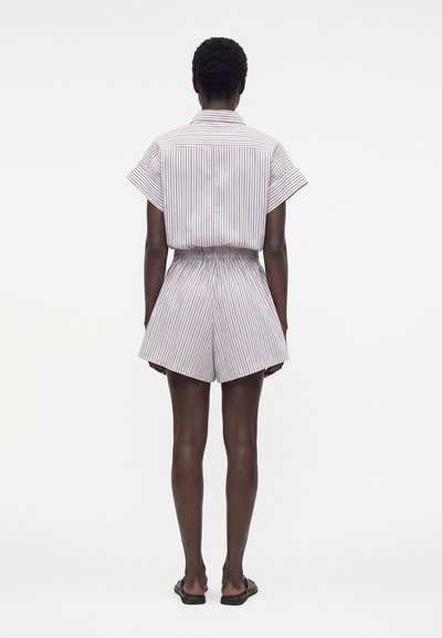 Άτομο όρθιο με την πλάτη γυρισμένη, φοράει λευκό κοντομάνικο πουκάμισο και σορτς με λεπτές κάθετες κόκκινες και μπλε ρίγες, και μαύρα σανδάλια.