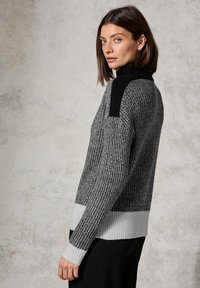 Pull en maille gris avec un design texturé, des accents noirs aux épaules et un ourlet côtelé blanc. Col montant, manches longues, coupe décontractée.