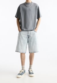 Camiseta gris de manga corta, pantalones cortos de mezclilla claros y zapatillas azules con suelas blancas. Presenta un corte relajado y cuello acanalado.