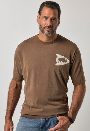 Mand med kort mørkt hår og skæg iført brun t-shirt med en hvid fiskelignende bøffel grafik på brystet og blå jeans.