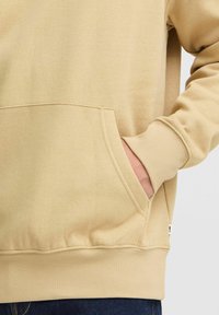 Sudadera beige con bolsillo frontal, puños acanalados y cinturilla. Tejido suave con un diseño minimalista y sutiles detalles de costura.
