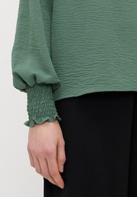 Roheline tekstuuriga bluus puff-särkidega; mansettidel smock-detailid ja volangservad. Sobib musta kangaga.