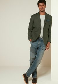 Dunkelgrüner Blazer mit einem grauen T-Shirt, blauen Jeans und braunen Wildlederschuhen. Der Blazer hat ein Einstecktuch; insgesamt entspannte Passform.