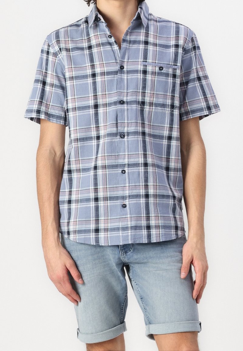 Homme portant une chemise à manches courtes à carreaux bleus avec poche poitrine et un short en jean bleu clair retroussé.