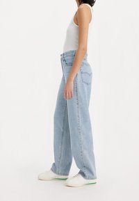 Personne portant un jean large bleu clair, un débardeur blanc et des baskets blanches, debout de profil devant un fond uni.