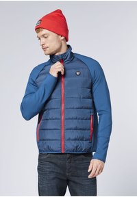 Blaue gesteppte Jacke mit Reißverschluss vorne, roten Akzenten und einem Logo-Abzeichen. Lange blaue Ärmel, zwei Seitentaschen und hoher Kragen. Kombiniert mit einer roten Beanie.