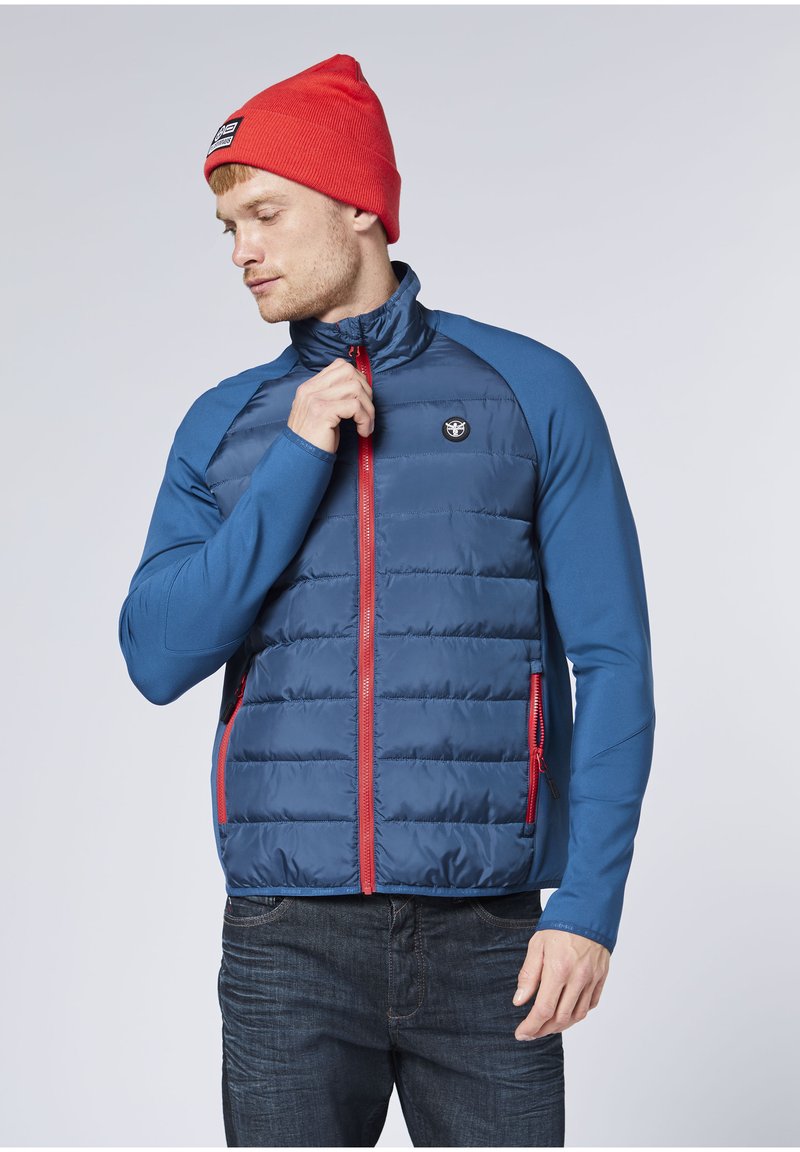 Blaue gesteppte Jacke mit Reißverschluss vorne, roten Akzenten und einem Logo-Abzeichen. Lange blaue Ärmel, zwei Seitentaschen und hoher Kragen. Kombiniert mit einer roten Beanie.