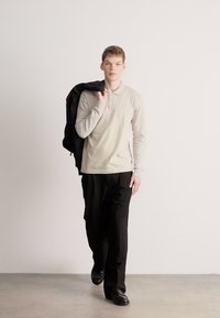 Svetlo siva polo majica z dolgimi rokavi, črne hlače in črni čevlji. Model nosi črno jakno čez ramo, pozira v minimalističnem okolju.