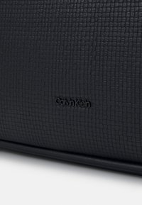 Surface en cuir noir texturé avec un petit motif en grille et le logo Calvin Klein embossé au centre.