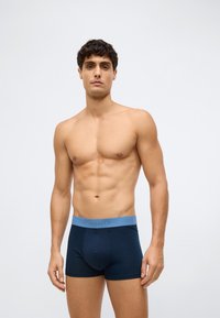 Fit young man wearing navy boxer briefs with a light blue waistband, standing against a plain light gray background.
