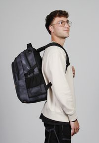 Mochila negra y gris con múltiples compartimentos, correas acolchadas y detalles de malla, llevada en la espalda de una persona, con un diseño elegante y moderno.