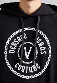 Felpa nera con cappuccio caratterizzata da una grande grafica rotonda bianca con la scritta "VERSACE JEANS COUTURE" e una "V" centrale, accentuata da un motivo a catena.